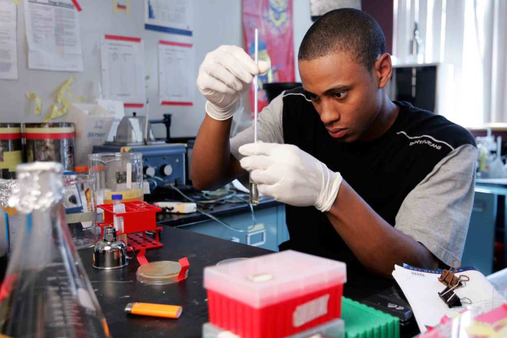 A Black student wearing white latex gloves holds a test tube and focuses intently on a pipette. They are in a chemistry lab surrounded by lab equipment.