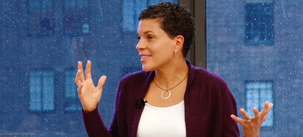 Michelle Alexander is speaking and gesturing with her hands. Her hair is short and she wears silver jewelry, a purple sweater, and white shirt.