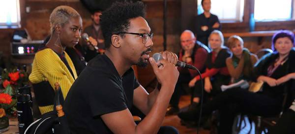 A seated Black person with a moustache and beard wearing a black t-shirt and eyeglasses speaks into a microphone before a seated crowd at the Summit on Creativity and Free Expression in Utah.