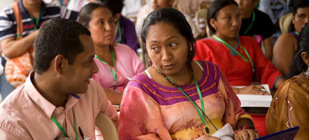 Indigenous activists in Peru sit at an oudoor conference wearing green lanyards. Two people sit in the foreground, one in a pink polo and short hair, another with their long hair tied back in a pink and yellow dress trimmed with maroon and orange on the neckline.