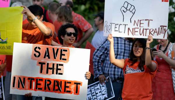Two people wearing orange stand outside with a crowd of protesters, holding protest signs. One with short curly hair and sunglasses holds a sign that reads "Save the Internet". Another wiith long dark hair holds a sign over their head with a hand drawn fist that reads "Protect Net Neutrality".