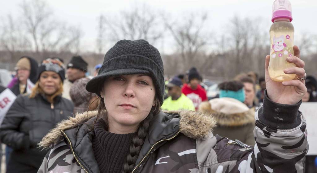 A Flint, Michigan protester wearing winter clothes holds a baby bottle full of dirty water. They have their hair in a long dark braid and a facial piercing beneath their left eye.