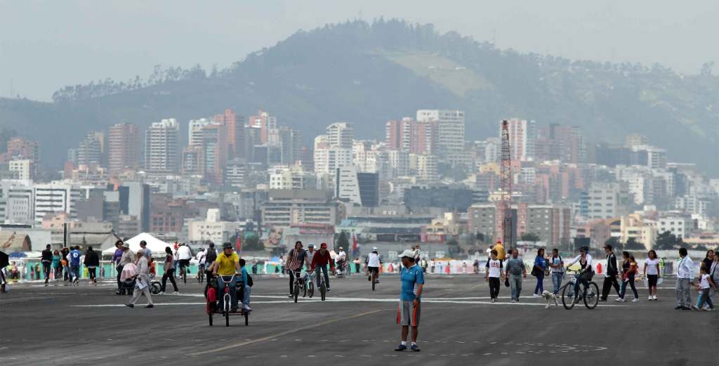 People walk across the pavement of an airport in Ecuador. Quito's cityscape is in the background.