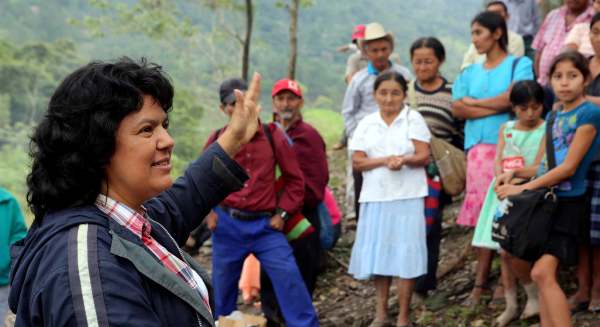 Berta Cáceres, smiling and waving a hand, speaks to community members about Aqua Zarca vote. They are outside, standing on dirt and surrounded by green trees.