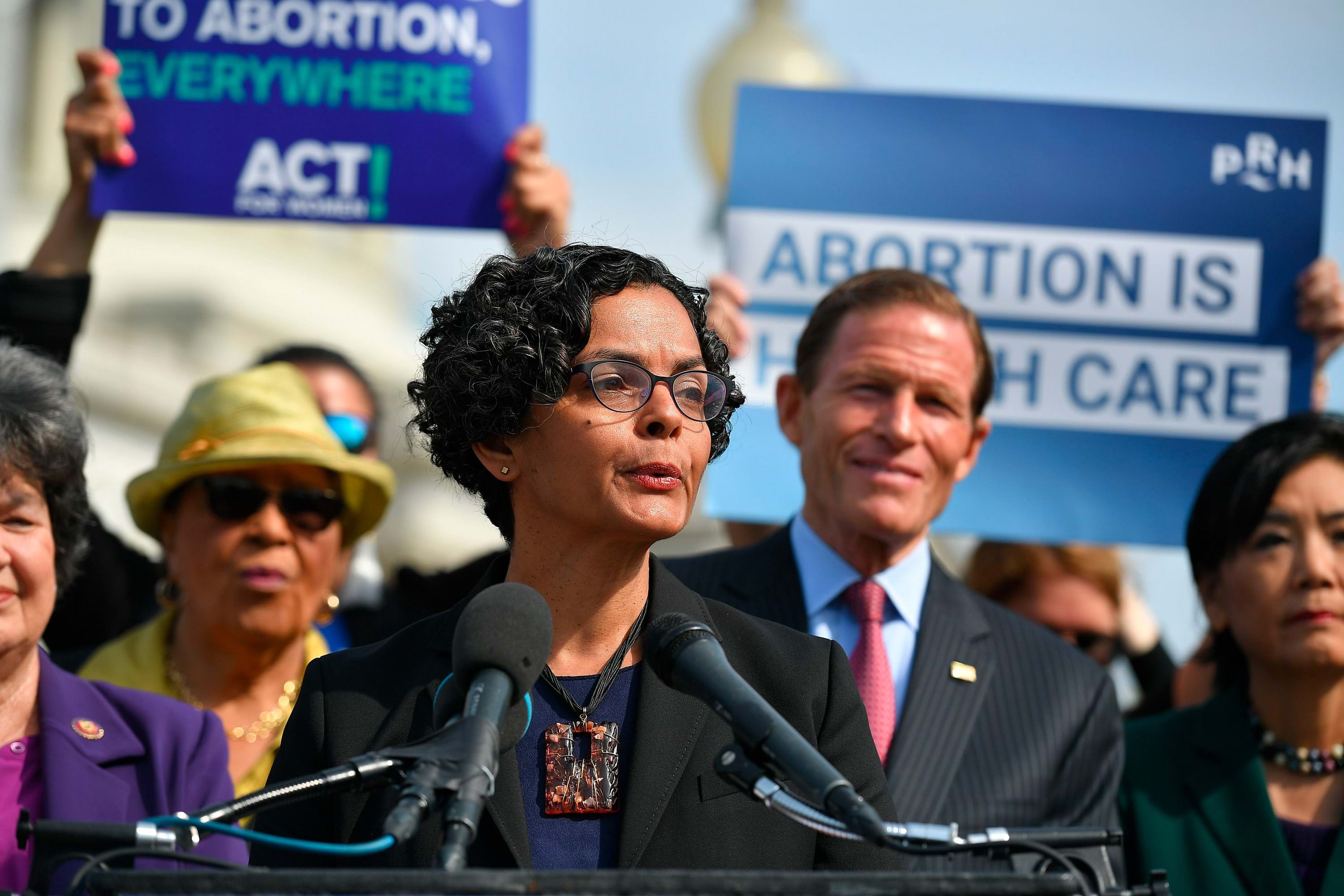 Lourdes Rivera speaks at a podium with people holding signs that read "Abortion is Health Care"