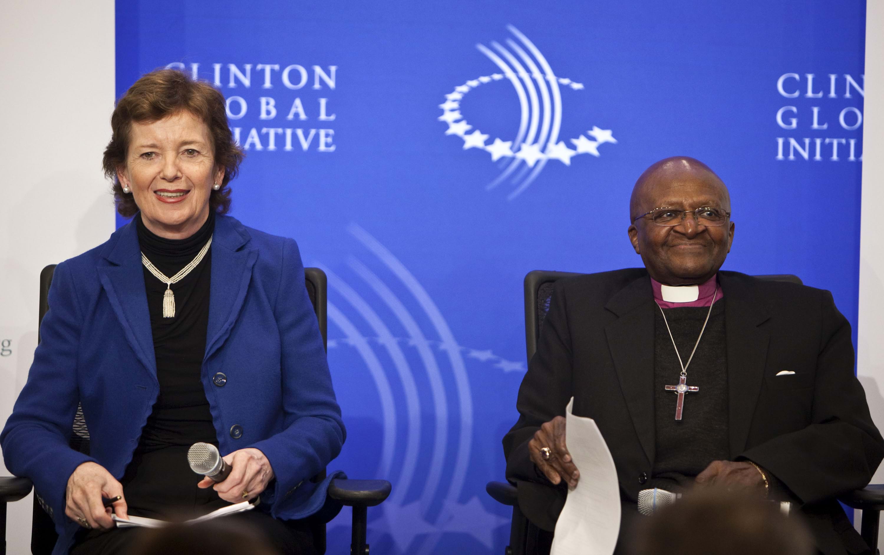 Mary Robinson and Desmond Tutu sit next to each other in front of a blue backdrop that has the Clinton Global Initiative logo on it.