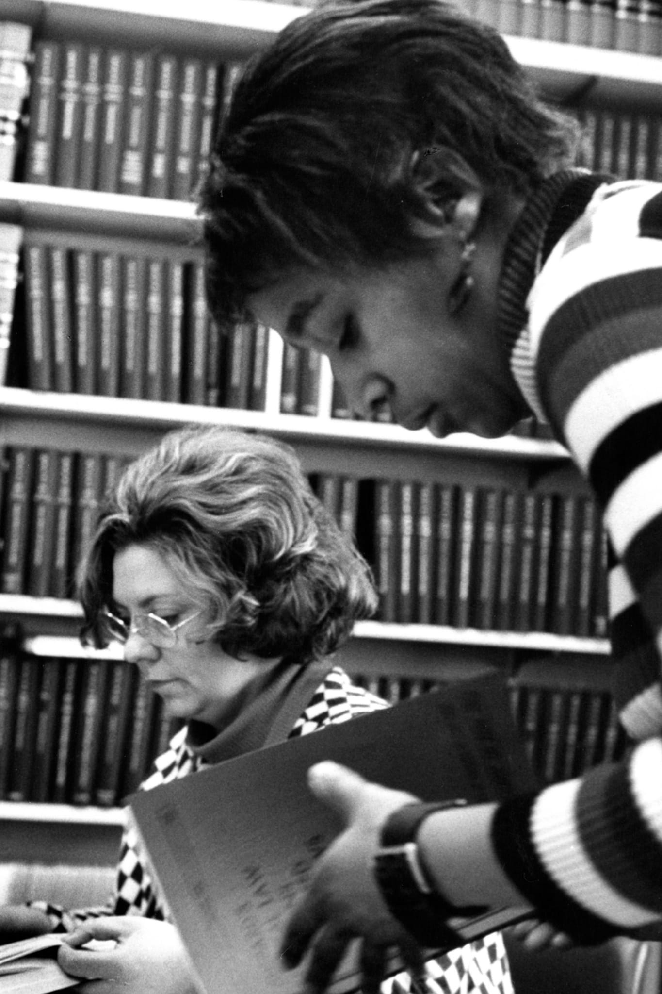 A black and white phot of a Black woman and white woman reading books in a library.