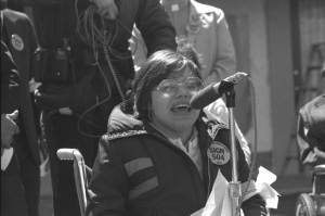 Judy Heumann in a wheelchair speaking into a microphone. She is wearing glasses and a coat with a Sign 504 sticker.