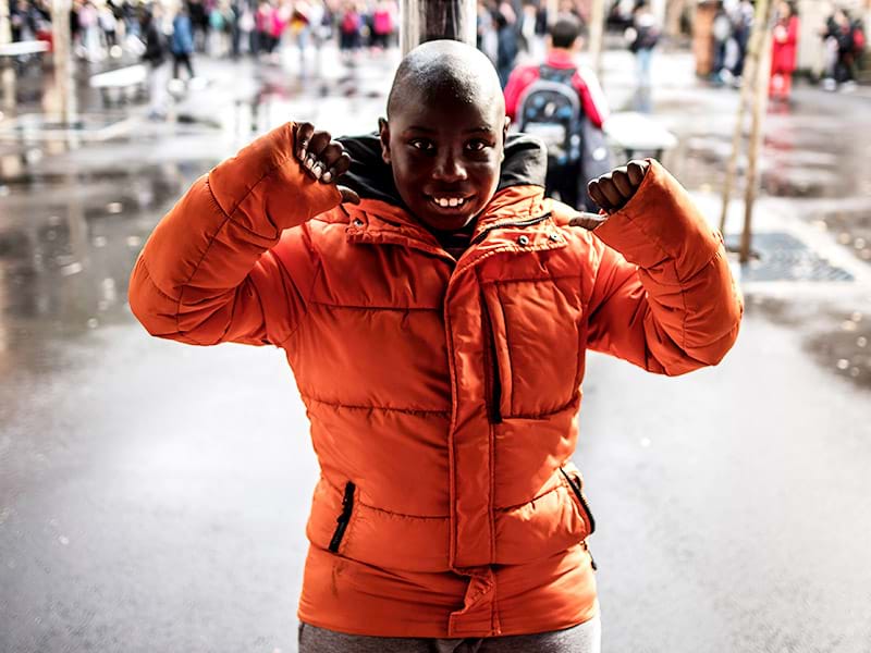 A young black boy with autism smiles with his arms raised in the schoolyard. He is wearing a bright orange puffy coat.
