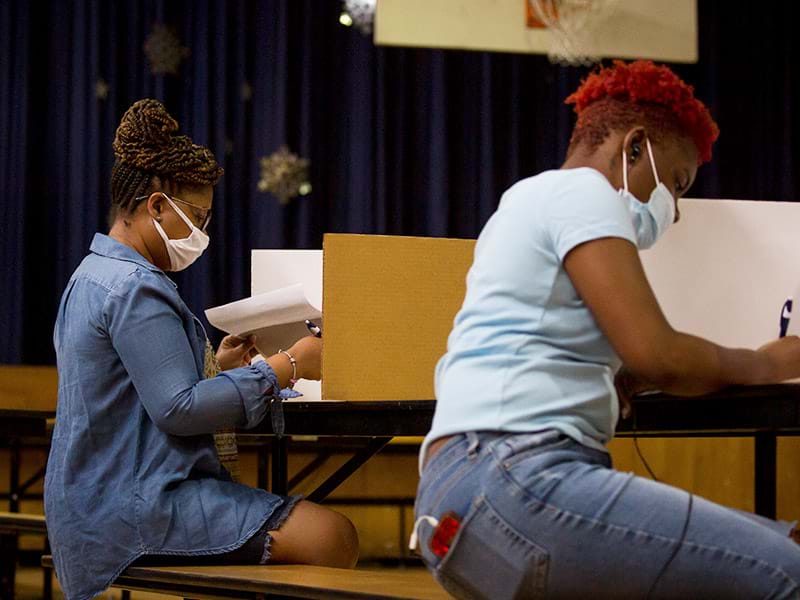 Two black women wearing protective masks and seated in a school gymnasium filling out ballots to vote.