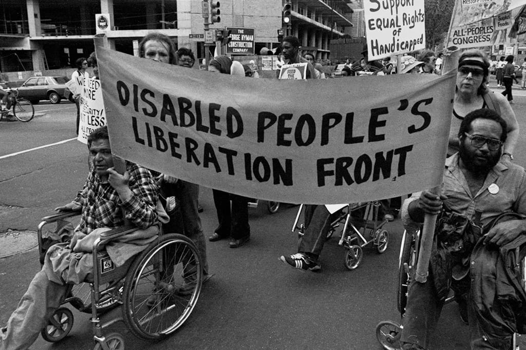 Two people in wheelchairs lead a protest with a banner that reads “Disabled People’s Liberation Front” with a crowd behind them.