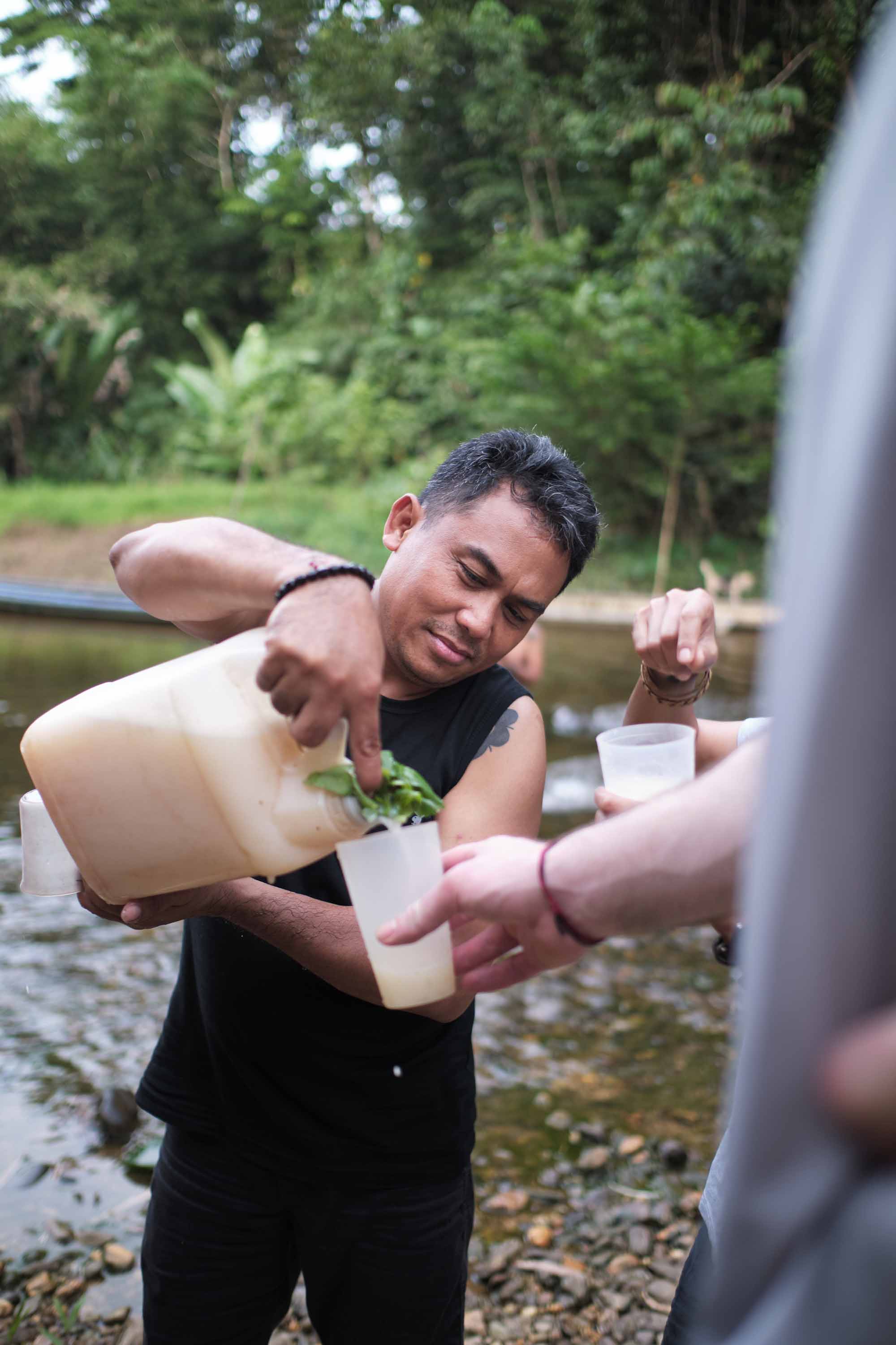 A man standing in a stream in black shirt and jeans pours a white drink from a jug to a hand holding a cup