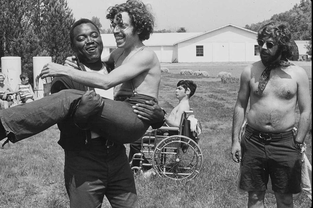 B&W photo of a Crip Camp counselor carrying one of the campers as another counselor looks on.