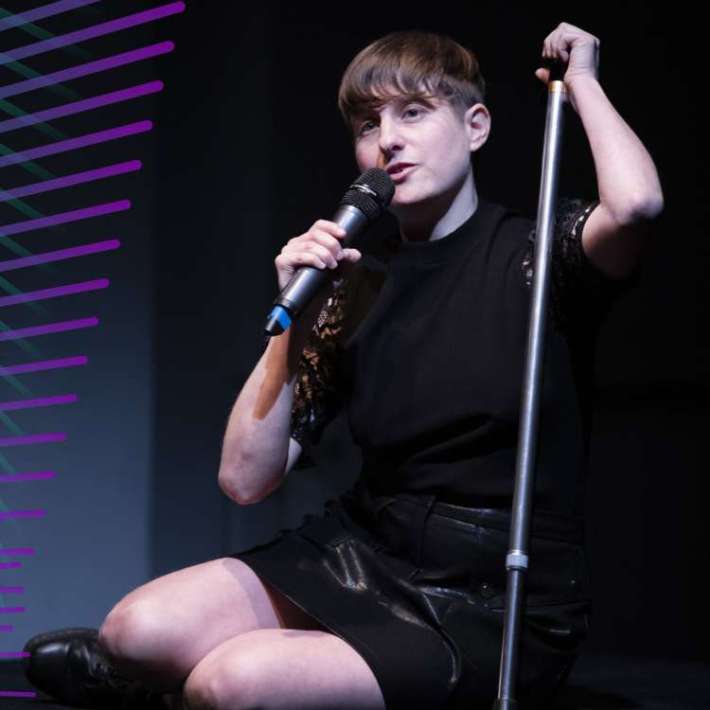 A white nonbinary person with styled short hair poses on stage. They're seated on the floor with their legs folded under them. They lean on their silver cane with their left hand and hold a microphone with their right hand. They wear a black top tucked into a sleek black skirt with black boots.