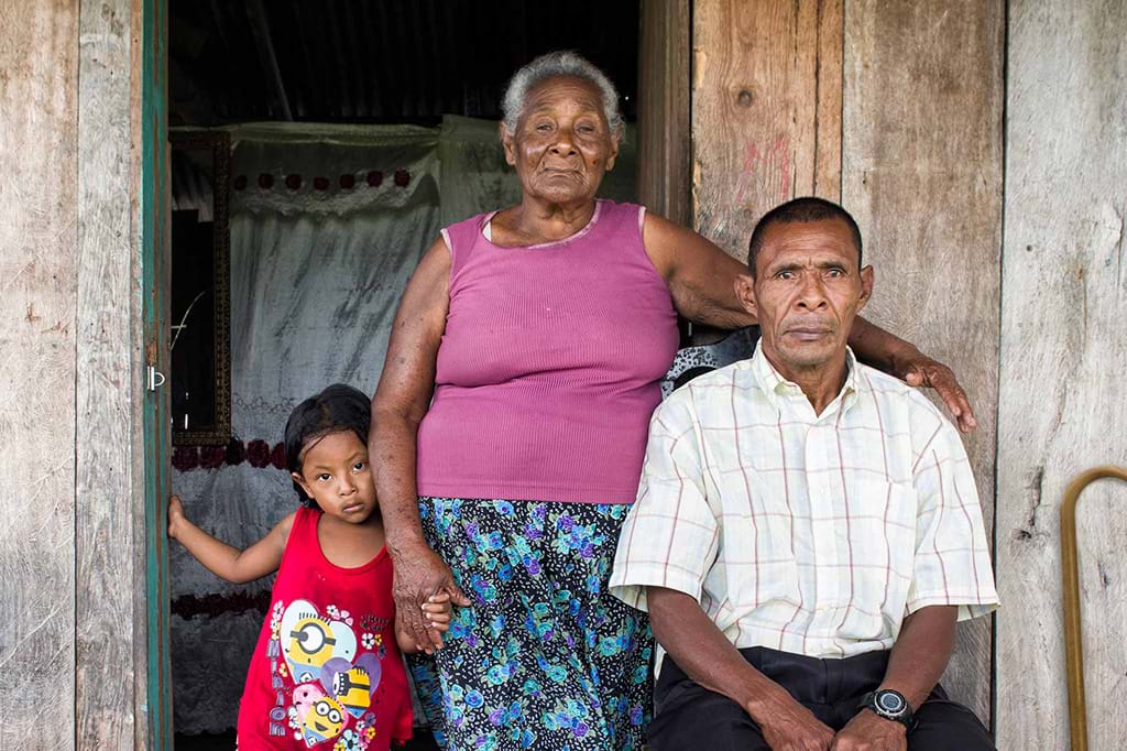A Honduran man sits in front of a wooden house surrounded by an older woman and a child. There is a cane leaning against the wall behind the man.  Photo: Antonio Busiello