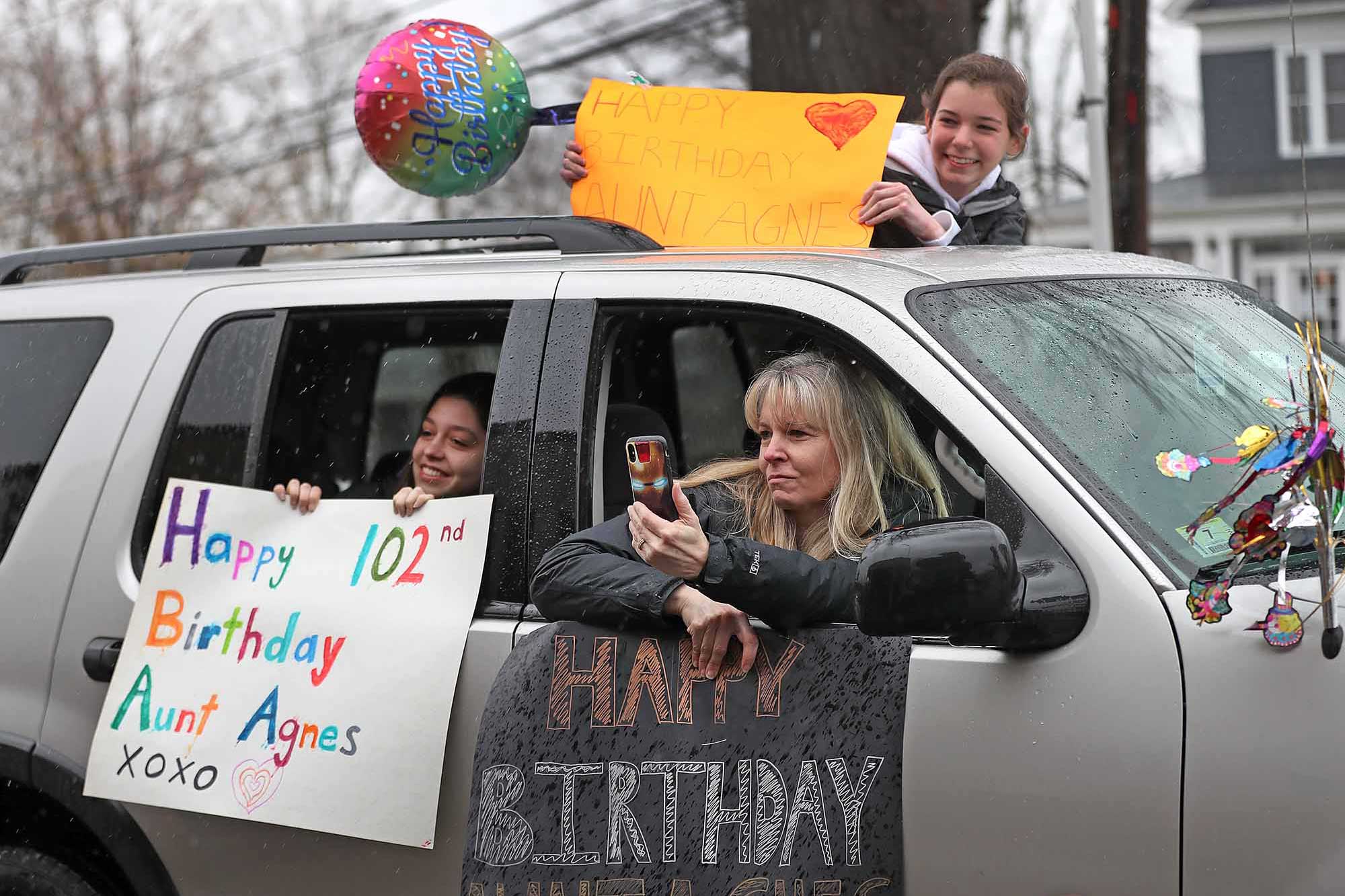  Three people hold colorful birthday signs outside of a car window.