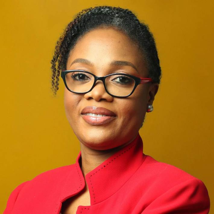 Portrait Olufunke Baruwa wearing a red top and black frame glasses against a mustard yellow background.