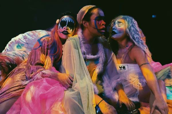 Three actors in colorful flowing garments sit next to one another and lean against one other