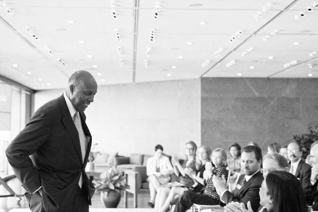 Vernon Jordan in a dark suit smiles towards a crowd with his hands in his pocket as the audience applauds him in a sunny room.