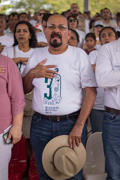 Marcedonio Cortave has his right hand on his chest while standing amongst community members. He wears belted blue jeans with a tucked in white graphic shirt that reads: “30 años Reserva de Biosfera Maya”. He holds a wide brimmed hat in his left hand.