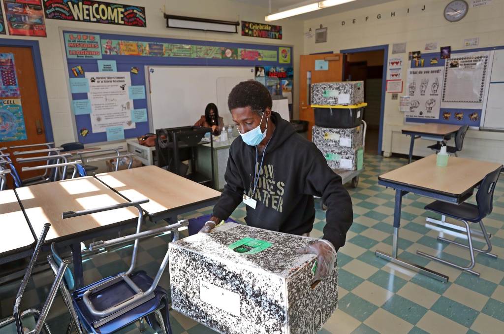 A Black man wearing a protective mask, a black sweatshirt that reads “our voices matter”, and medical gloves carries a box in a classroom while a woman sits as a desk in the background.