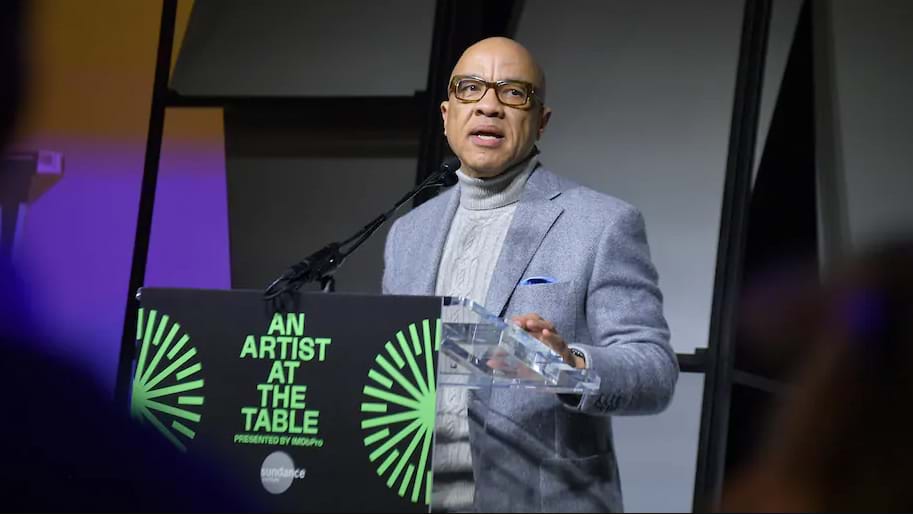 Darren Walker speaking at the 'An Artist at the Table' event is wearing a gray blazer and light gray button down shirt and dark rim glasses.
