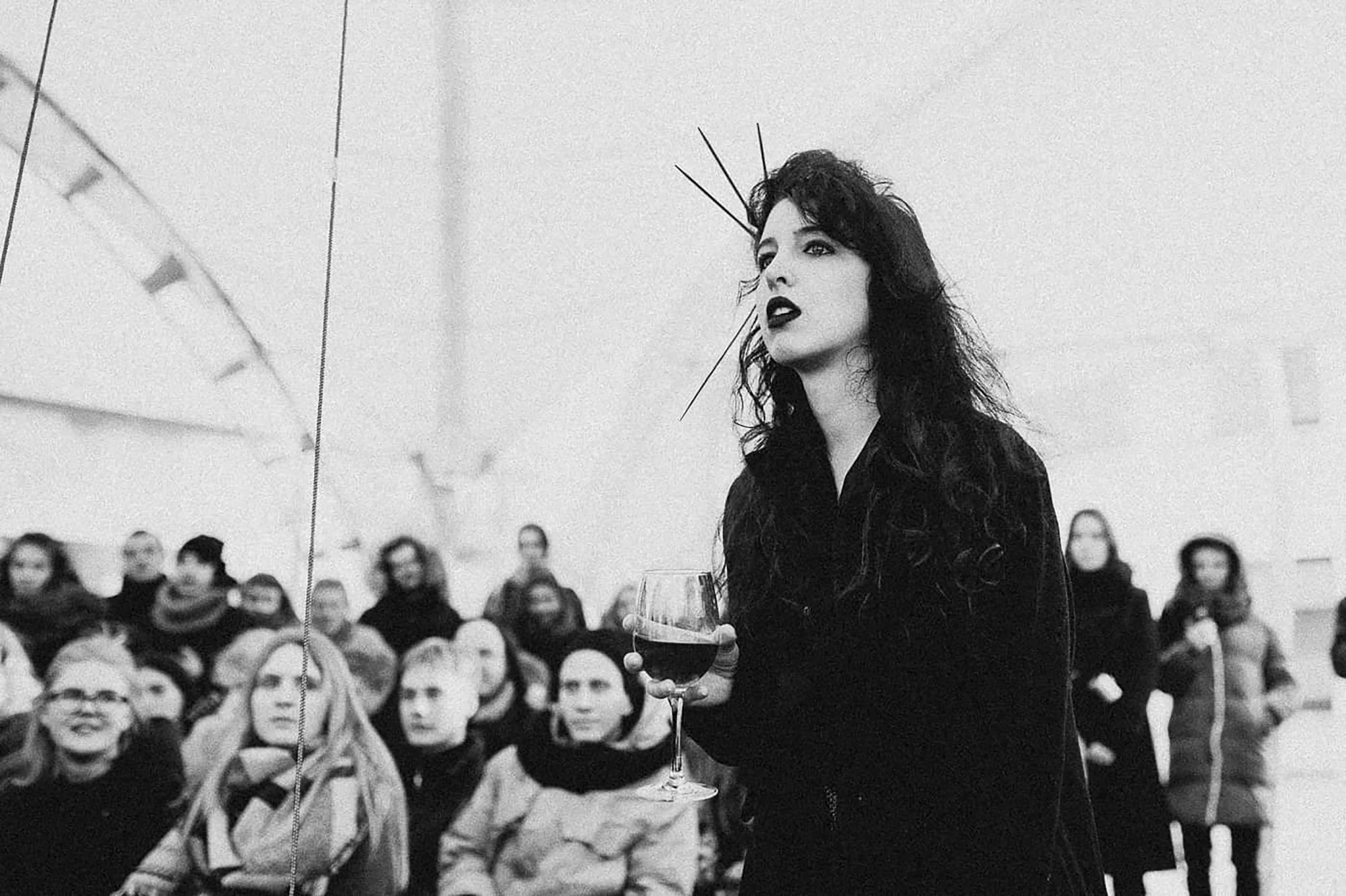 A black-and-white image of a woman, Khawla Ibraheem, standing in a dark outfit with shoulder-length dark hair. She has her back to an audience watching her, and small spikes coming out of one side of her head.