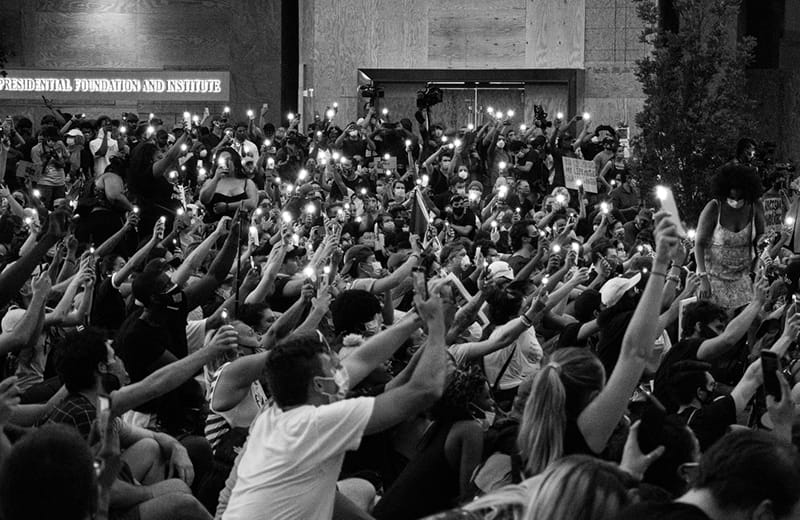 B&W image of a group of people holding their mobile devices in front of them with the flashlight feature on.