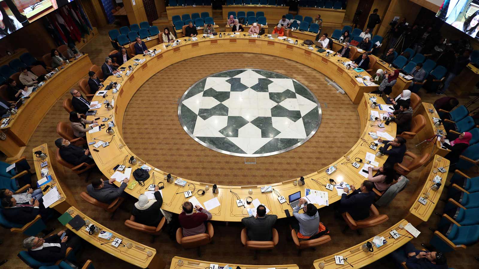 A view of a circular conference table at the United Nations with speakers seated in front of microphones. At the center is a mosaic of green and white marble.