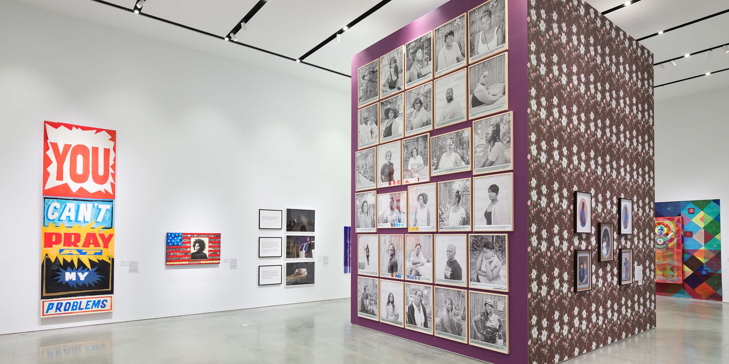 In a bright white gallery, a purple ceiling height structure is hung with a grid of black and white portraits. Around the gallery are colorful paintings and photographs.