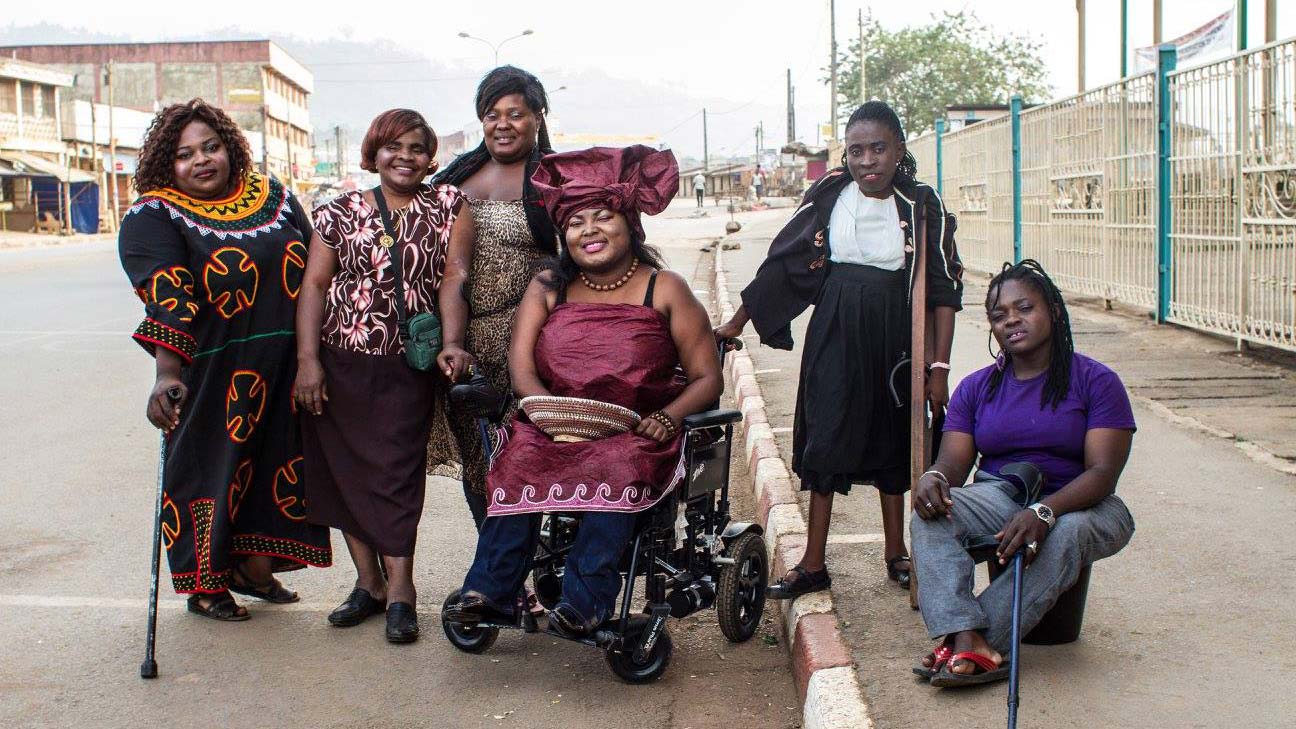 On a city street, six Black women with disabilities dressed in vibrant prints and colors, smile together. The women at the center uses a powerchair while others around her use canes and crutches.