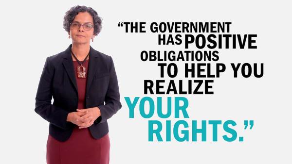 Lourdes Rivera has short dark curly hair and is wearing dark-rimmed glasses, earrings with an orange stone, a black blazer over a burgundy dress, and a large pendant necklace. Next to her is copy that reads, "The Government has positive obligations to help you realize your rights."