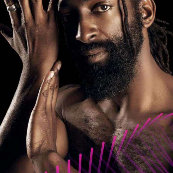 An African-American and Indigenous person with dark chocolate skin from his mother. He has almond-shaped eyes with long lashes, long black locs tied in a low braid with a single loc visible across his cheek, and a full beard. He is bare-chested with his left hand raised artistically near his face.