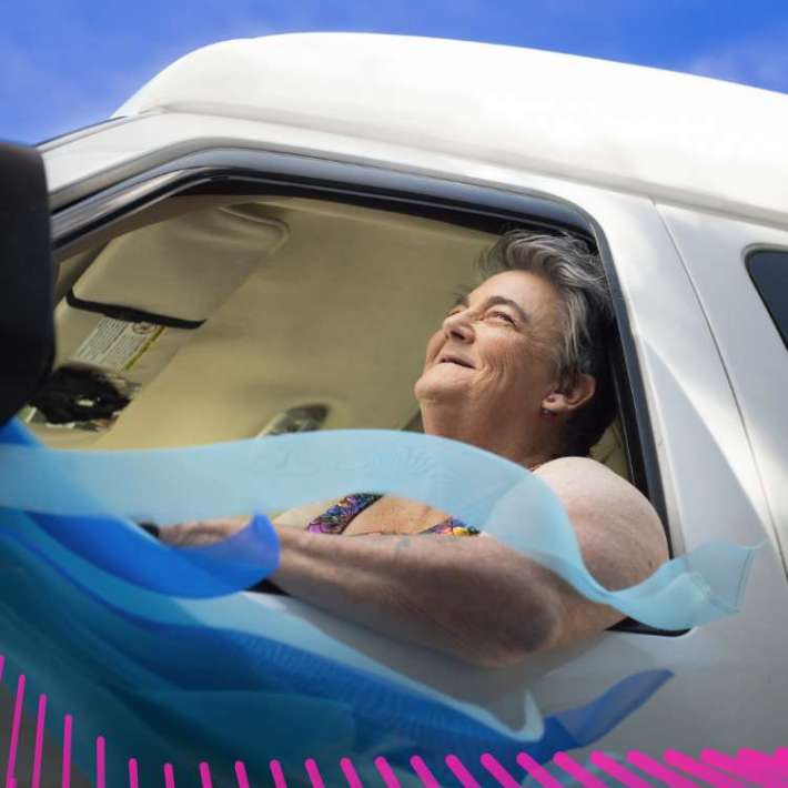 An older, large, white woman with short, greying hair, sitting and smiling in the driver seat of a white van. She is wearing a colorful patterned sleeveless top. In front of her are sky blue streamers attached to the side-view mirror of the van that flow and curl in the wind and cover part of her torso.