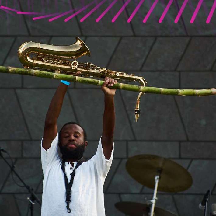 A Black man holding a tenor saxophone and a stalk of sugar cane above his head. He is wearing a white, short-sleeved shirt and a black saxophone neckstrap. Behind him is a wall of gray stone, cymbals, and microphones on stands.