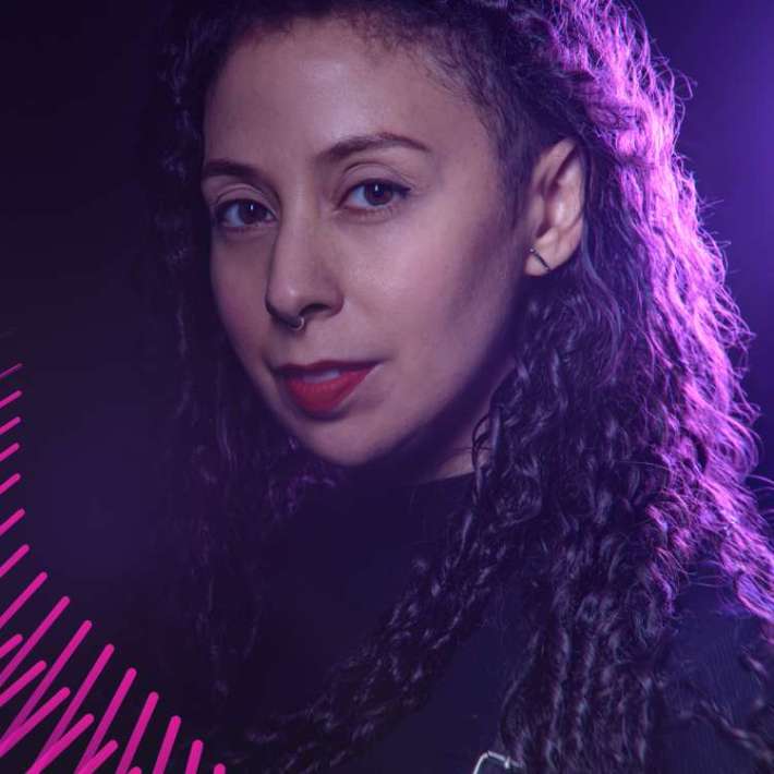 A multi-heritage woman with long tightly wound dark brown locks, wearing red lipstick and a high neck black shirt. Background is darkly lit with one purple light illuminating her left side onto her hair.