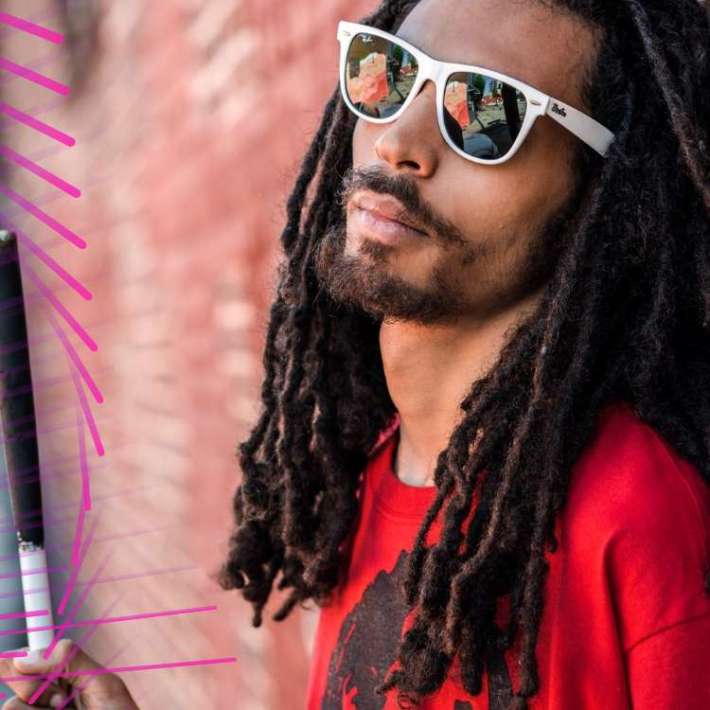 A biracial man with dreadlocks. He is wearing a red shirt and sunglasses and stands beside a brick wall, holding a white cane.
