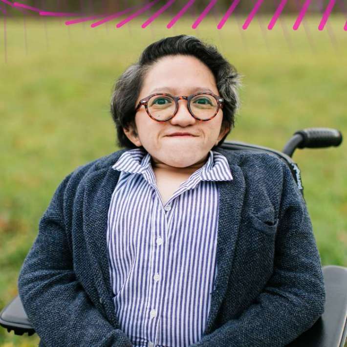 Photo of Ho, an Asian American woman. She is wearing a striped shirt, a dark blue cardigan, and red glasses. She sits in a power wheelchair in a field.