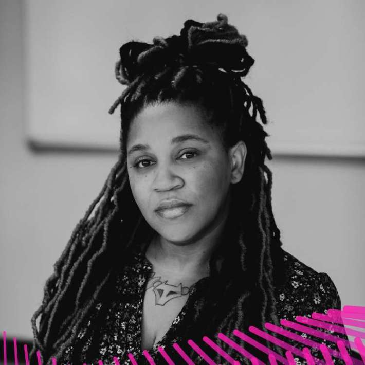 A Black woman with faux locs in a half up/down ponytail hairstyle. She is wearing a blouse patterned with tulips and staring into the camera.]