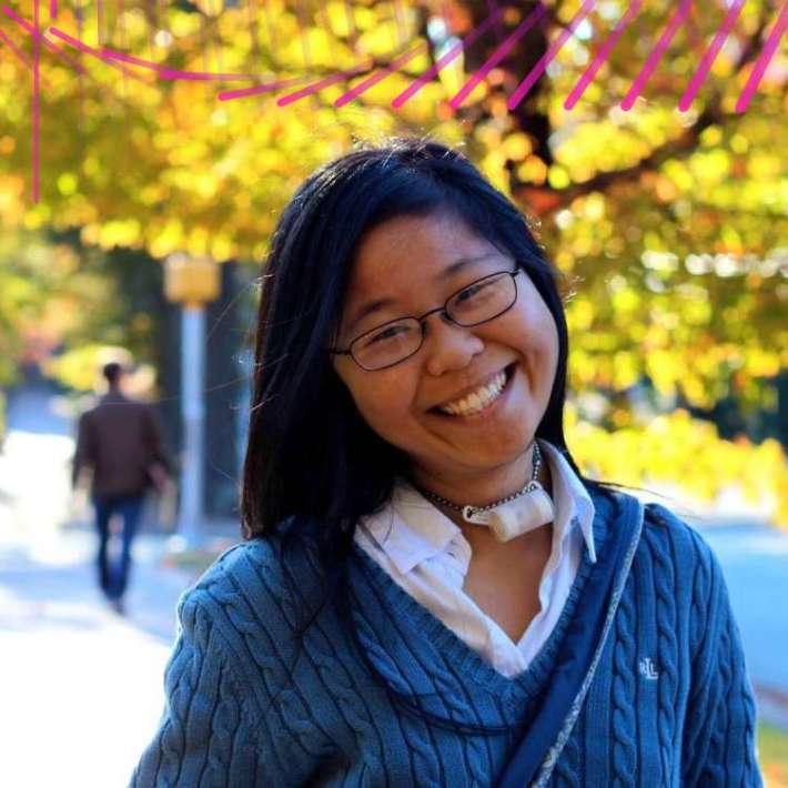 Lu, a Chinese American woman, smiling warmly at the camera. She is wearing black-framed glasses, a tracheostomy tube, and a blue sweater over a white button-down shirt. She is standing in front of a tree with yellow leaves.