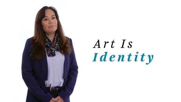 Lori Pourier wears a blue suit over a light blue blouse and has a multicolored scarf around her neck. The phrase "Art Is Identity" appears to the right.