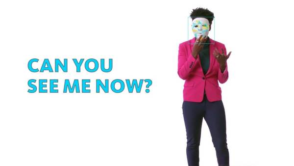 Joy Buolamwini is a Black person wearing a bright pink blazer and matching glasses. Joy holds a faceless white mask to her face. The phrase "Can you see me now?" appears to the left.