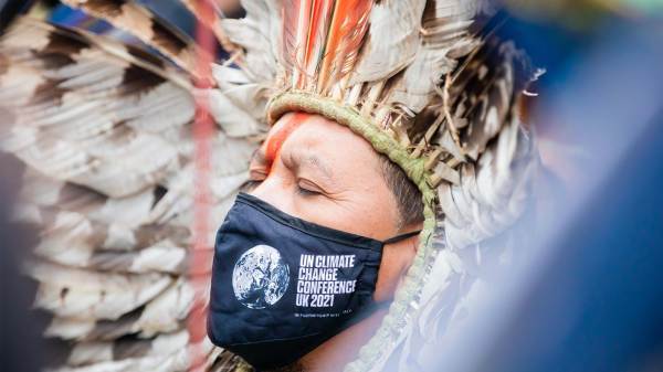 Indigenous man wearing traditional clothing at UN Climate Change Conference COP26 in Glasgow. He has his eyes closed and wearing a mask with COP26 branding. 