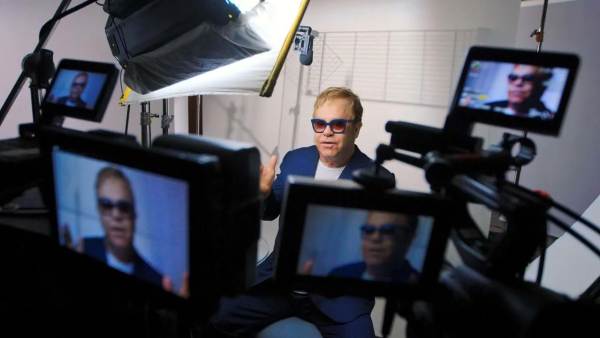 Elton John sitting on a film set in front of multiple cameras with his face displayed on all monitors. 