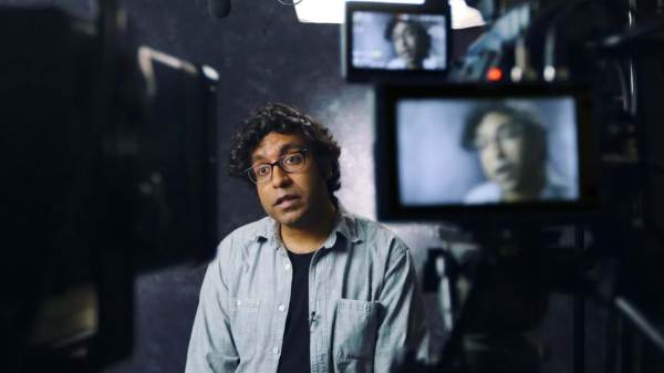 Hari Kondabolu sitting on a film set in front of multiple cameras with his face displayed on all monitors. 