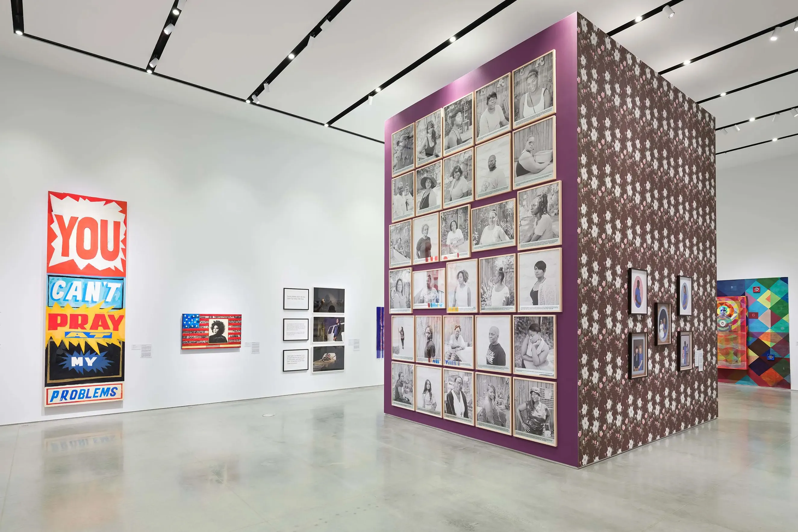 In a bright white gallery, a purple ceiling height structure is hung with a grid of black and white portraits. Around the gallery are colorful paintings and photographs.